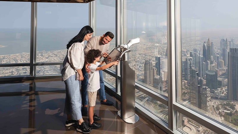 A family looks through a telescope at a cityscape from a high observation deck with large windows.